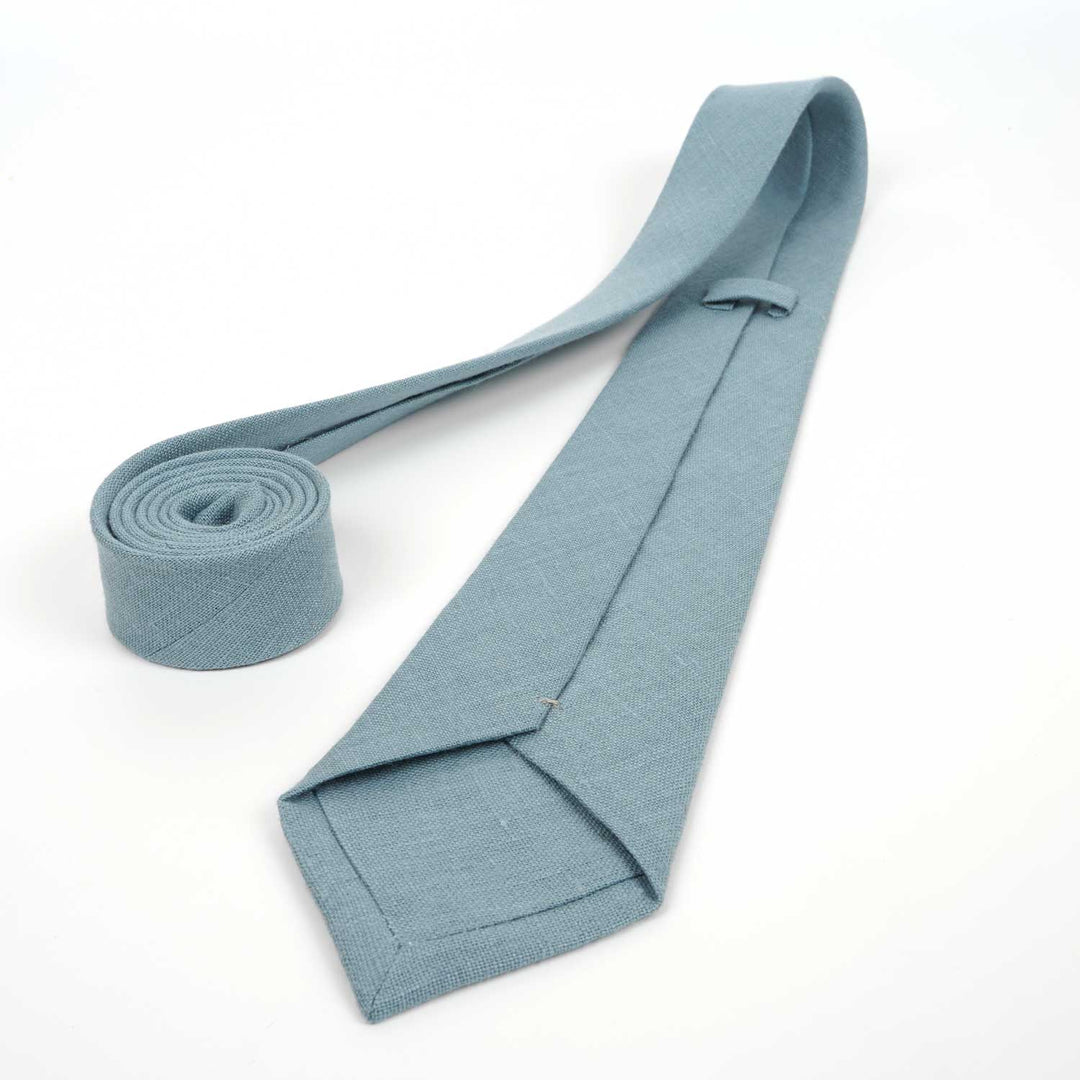 The reverse side of a dusty blue linen necktie shown upside down, highlighting the fabric lining, stitching, and keeper loop against a white background.