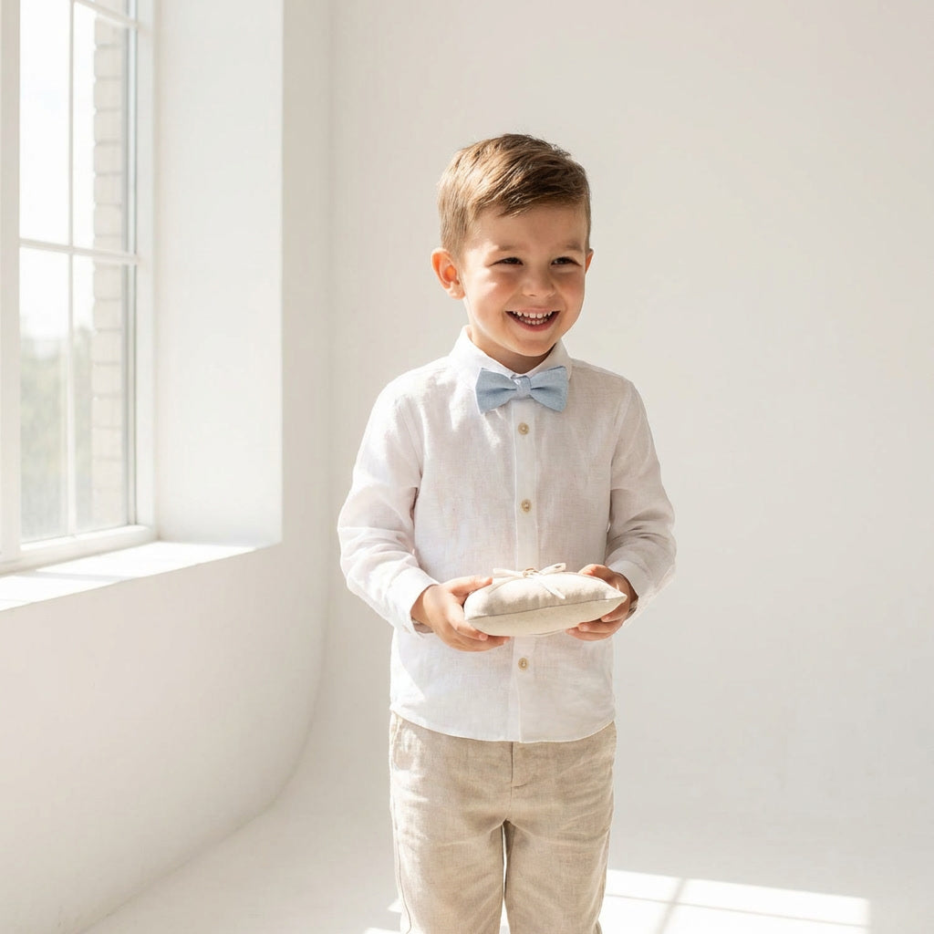 Young boy wearing a Hemmitie pale blue linen pre-tied bow tie with a white shirt and beige pants in a bright setting.