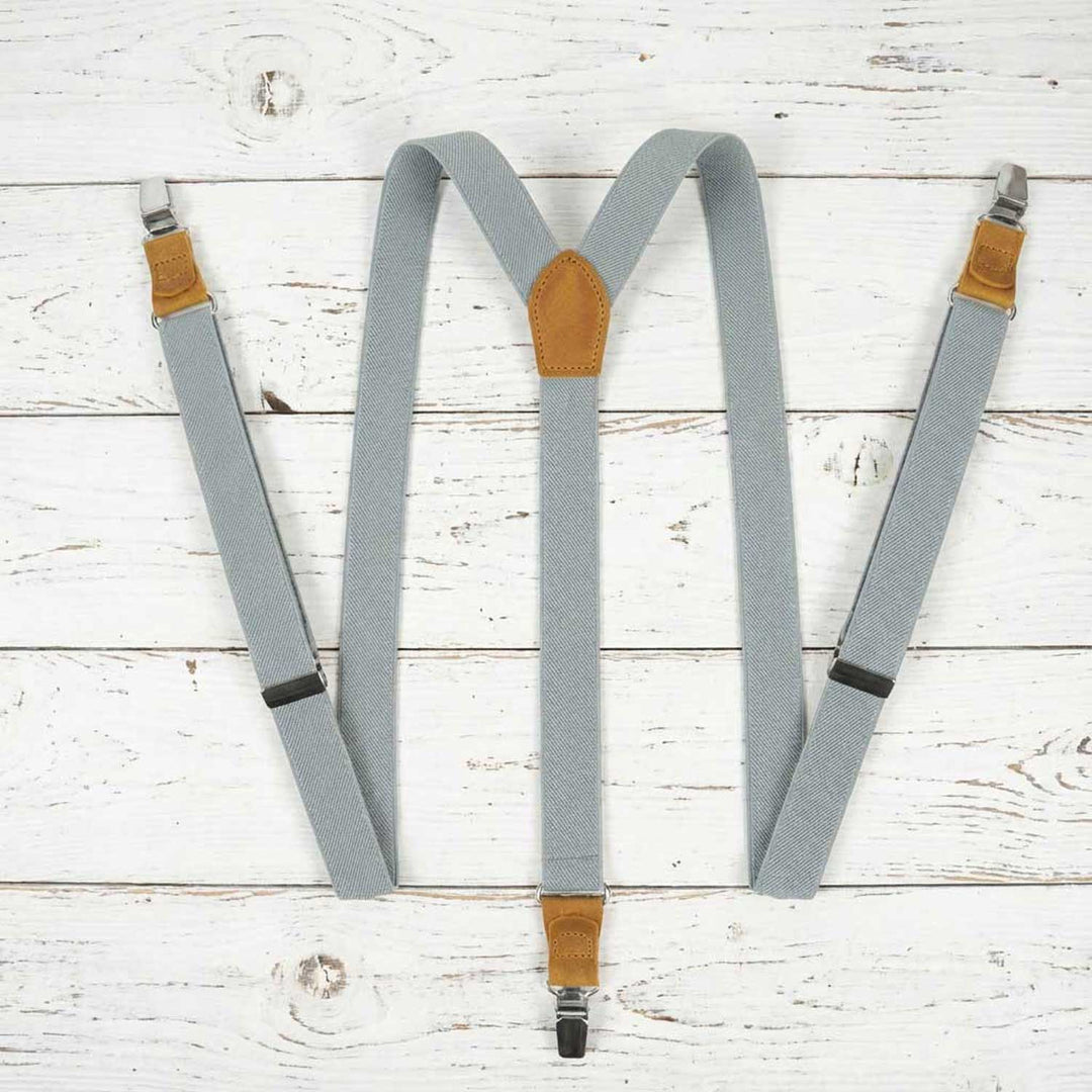 Top-down full view of light gray elastic Y-back suspenders with mustard natural leather accents, displayed on a rustic white wooden surface.
