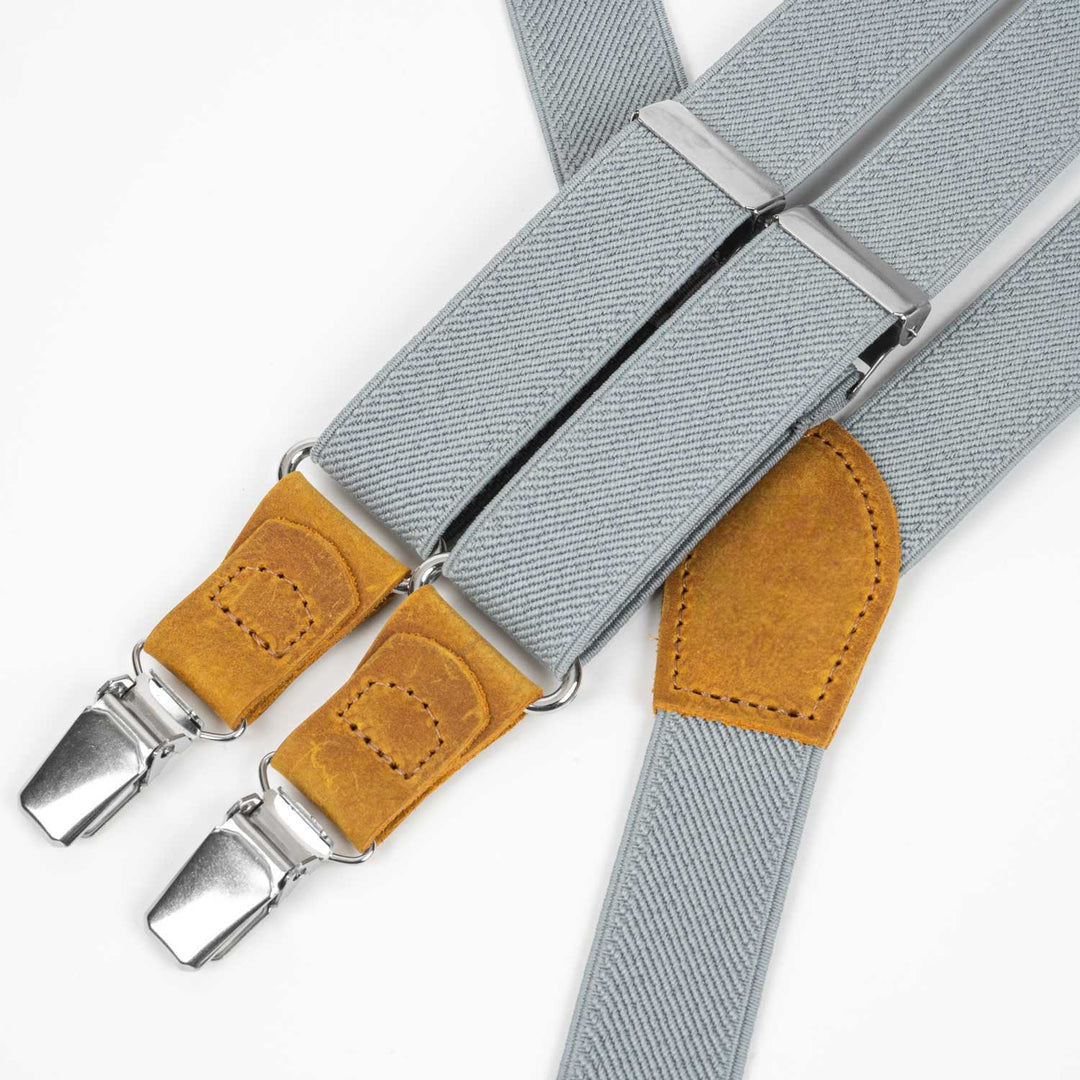 Light gray elastic Y-back suspenders with mustard yellow leather attachments and polished silver clips, laid flat on a clean white background.