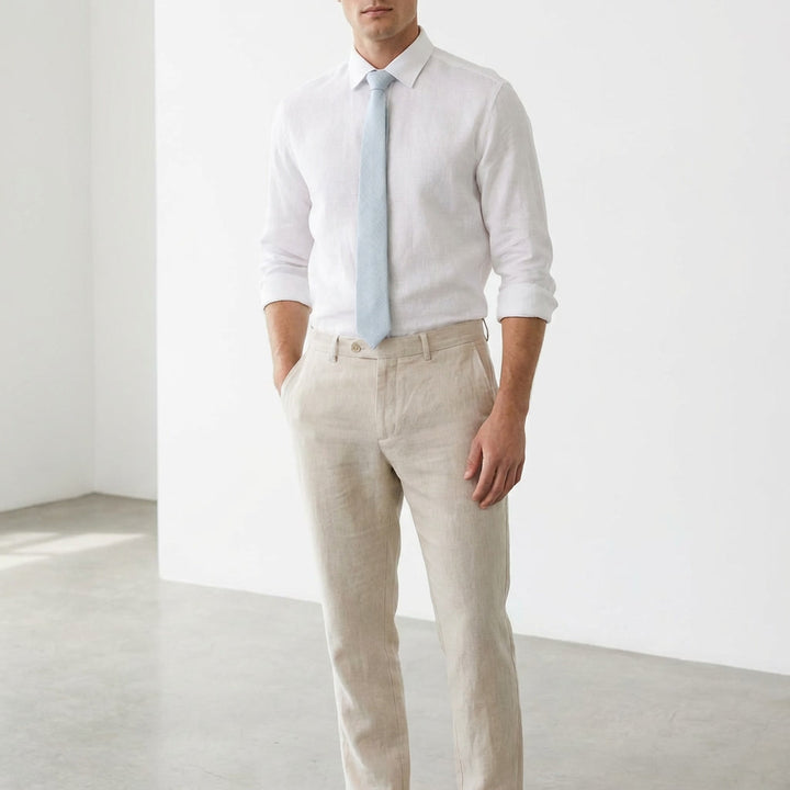 A man styled in a modern fit pale blue linen necktie and a matching white linen button-down shirt paired with beige trousers, set against a white studio background.