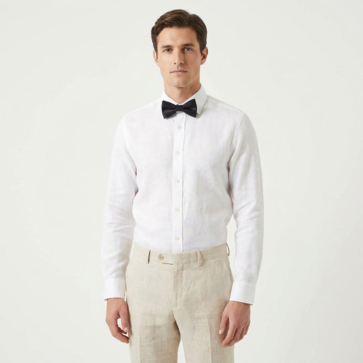 A man wearing a black pre-tied satin bow tie with a crisp white dress shirt and beige trousers, standing against a light gray background.