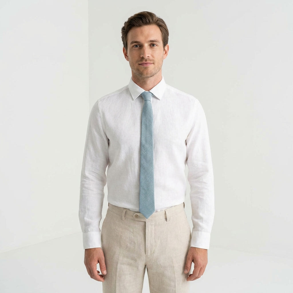 A man wearing a dusty blue linen necktie, a crisp white button-down shirt, and tailored beige trousers against a clean white background.