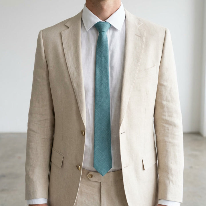 A man wearing a modern-fit beige suit and a crisp white shirt, accessorized with a textured teal blue linen necktie, posed against a soft neutral background.