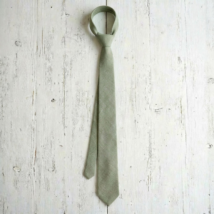 Top-down view of a sage green linen necktie, neatly tied and placed on a light-colored natural wood surface.